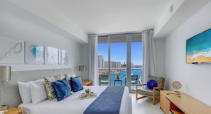 A modern hotel room with a large bed, a desk, a chair, and a view of a cityscape through a large window.