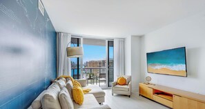 A modern living room with a sofa, armchair, and TV, a large window with a city view, and a wall with a blue design.