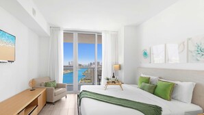Habitació d'hotel moderna amb un llit gran, un escriptori, una cadira i vista a la ciutat i al mar a través de grans finestres.