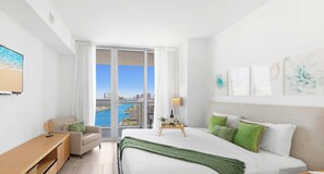 Quarto de hotel moderno com cama grande, escrivaninha, cadeira e vista para a cidade e para a água através de grandes janelas.