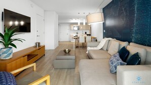 Ein modernes Wohnzimmer mit einer Couch, Hocker und Fernseher. Im Hintergrund ist eine Küche zu sehen und an der Wand hängt eine Karte.
