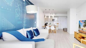 Una sala de estar moderna con un sofá blanco, cojines azules, una zona de comedor con una mesa y sillas blancas, una cocina con armarios blancos y un televisor sobre un mueble de madera.