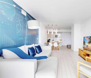 Un salon moderne avec un canapé blanc, des coussins bleus, un coin repas avec une table et des chaises blanches, une cuisine avec des meubles blancs et un téléviseur posé sur un meuble en bois.