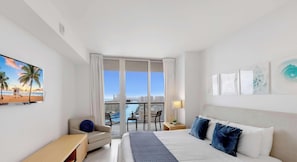 A hotel room with a bed, a desk, a chair, a TV, a balcony with a view of the city and a pool.