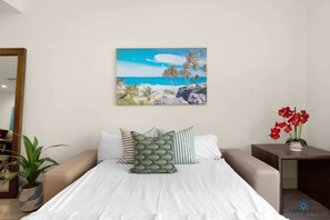 Une chambre à coucher avec un lit, des oreillers, un tableau représentant une scène de plage, une plante en pot et une petite table avec des fleurs.