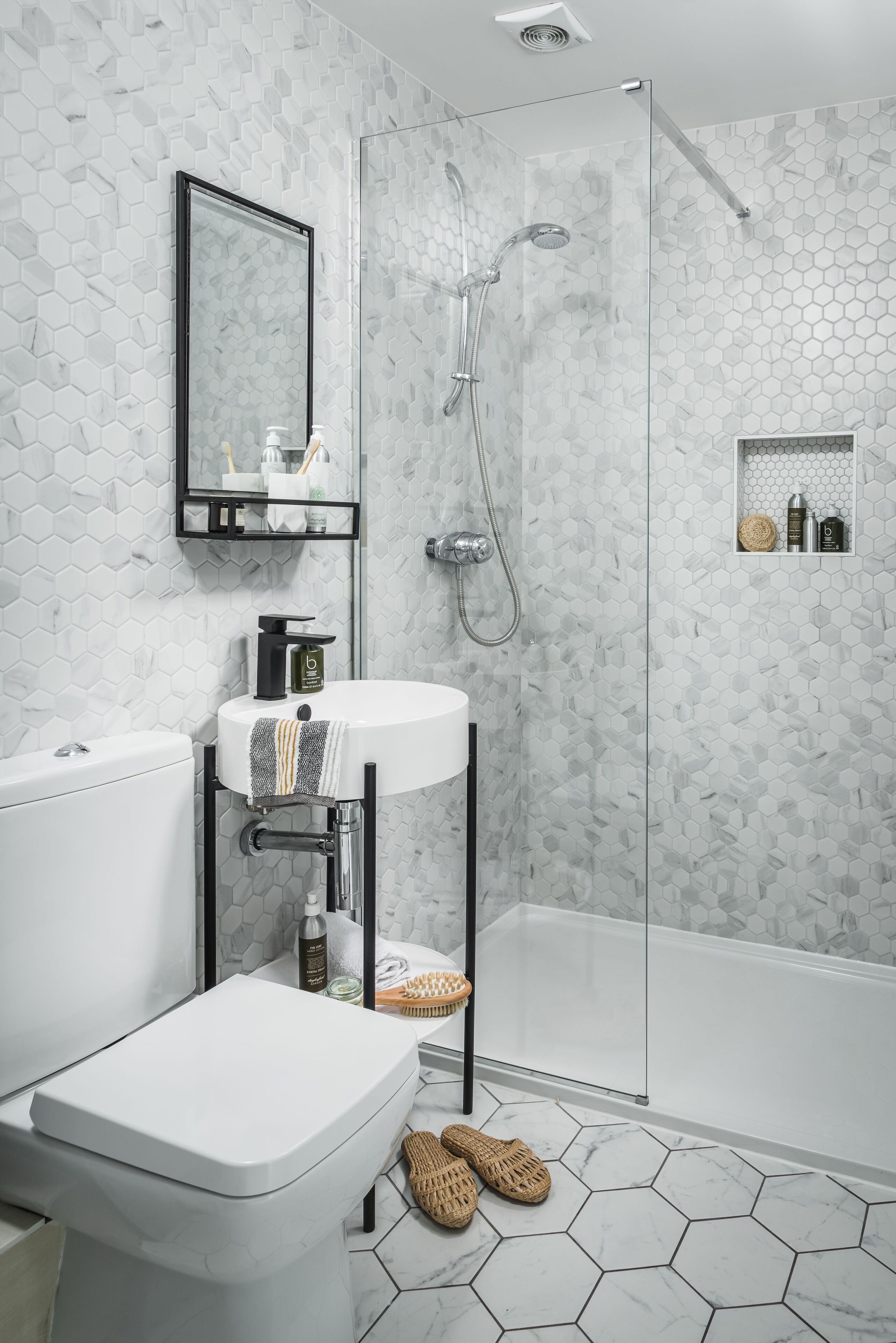 Un bagno moderno con pavimento in piastrelle esagonali, un water bianco, un lavandino con un asciugamano, uno spazio doccia in vetro e uno specchio sopra il lavandino.