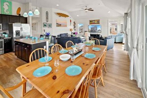 A modern kitchen with a central island, dining area with a wooden table set for four, and a living area with a sofa and TV.