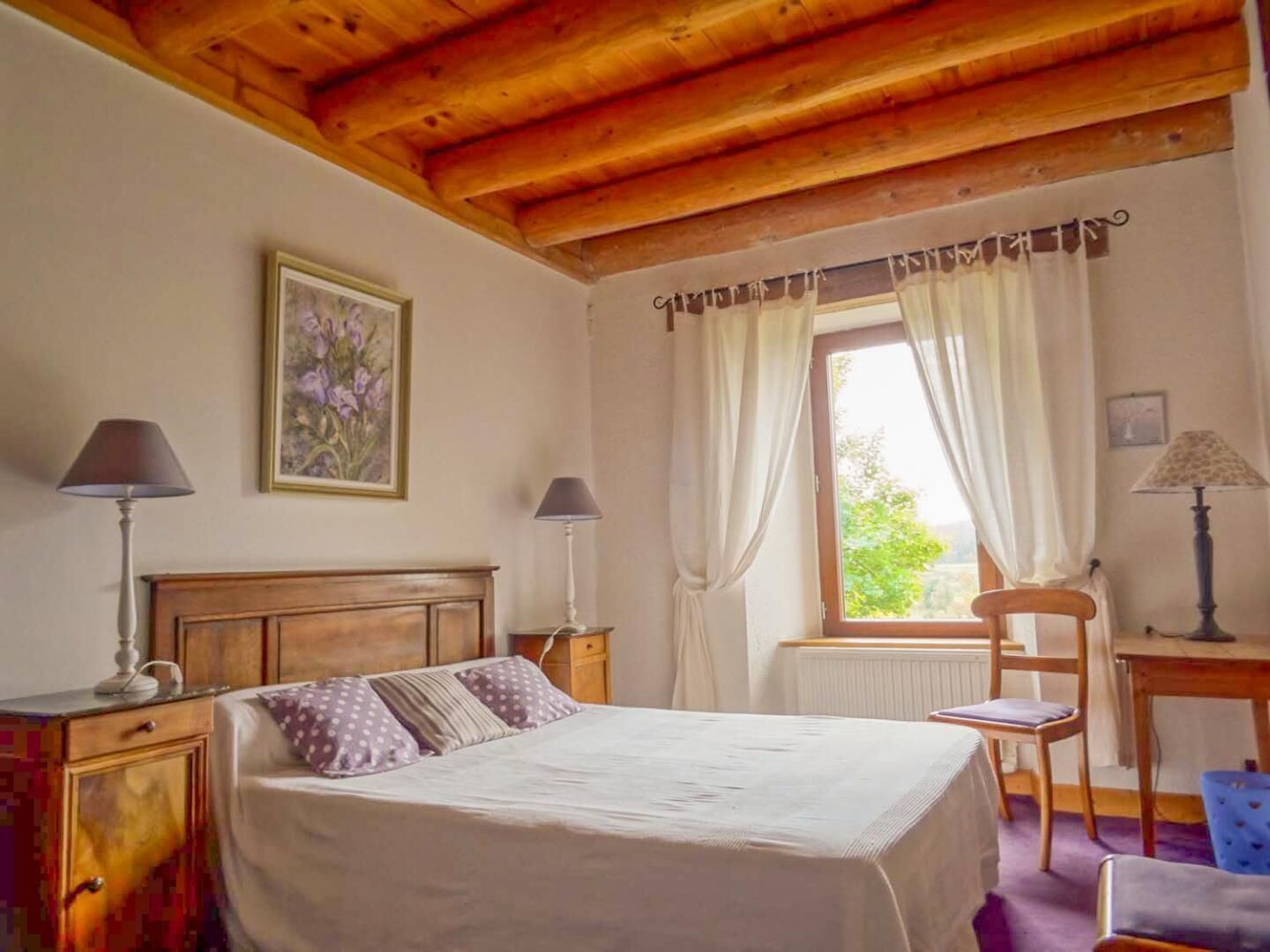 Un dormitorio con cabecera de madera, cama con ropa blanca, dos lámparas de noche, una ventana con cortinas, una silla y un escritorio con lámpara.