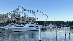 A marina with several yachts docked, a large metal structure in the background, and buildings across the water.