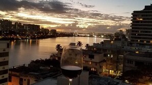 A glass of red wine on a balcony overlooking a cityscape at dusk with a body of water in the foreground.