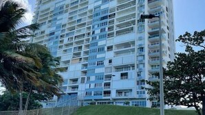 A high-rise building with multiple balconies, surrounded by greenery and a fenced area.
