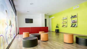 Un área de espera moderna con un banco rojo, varios otomanes redondos, un televisor de pantalla plana y una pared verde brillante con estanterías para revistas.