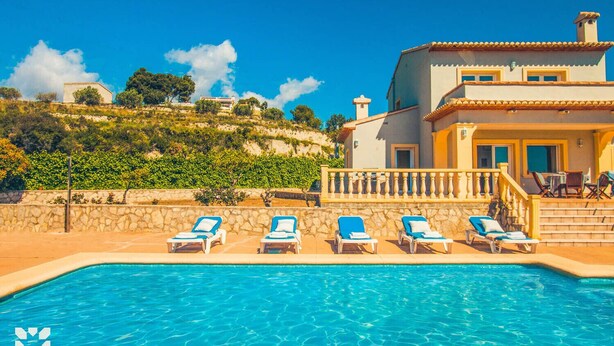 A swimming pool with lounge chairs, a stone wall, and a building with a balcony. - Villa AZALEA by Abahana Villas (Teulada)
