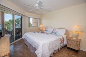 A bedroom with a large bed, two bedside lamps, a ceiling fan, and a view of the outdoors through a sliding glass door. - Easy Beach Living | Resort Pool & Spa (St. Pete Beach)
