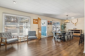 Une pièce spacieuse avec un plancher en bois, une table à manger avec des chaises et une grande fenêtre équipée de stores.