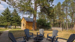 A wooden cabin with a metal roof, surrounded by a gravel area with four Adirondack chairs and a fire pit. - Blue Roof Lodge: Pet-Friendly, Walkable to Downtown Custer (Custer)