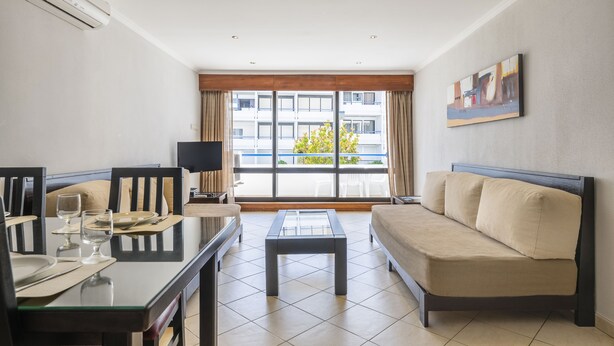 Ein modernes Wohnzimmer mit einem Glastisch, einem beigen Sofa und einem Essbereich mit Stühlen und einem Fernseher.
