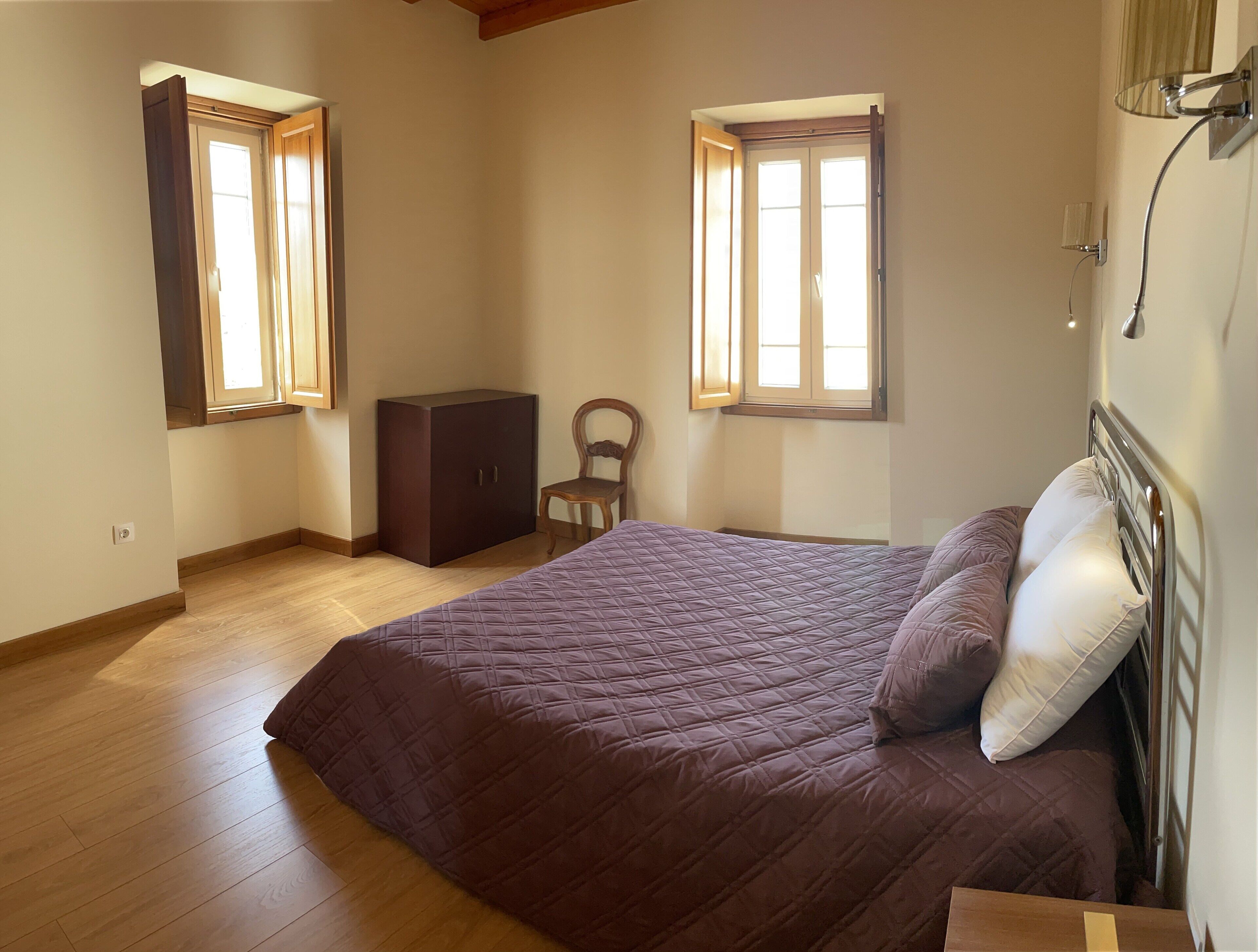 A bedroom with a bed, a wooden wardrobe, and two windows.