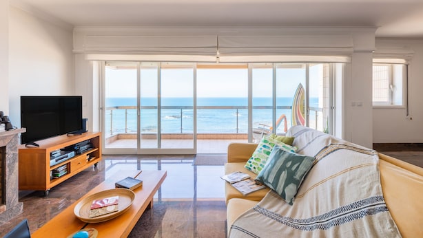 A spacious living room with a large TV, a wooden coffee table, and a sofa with colorful cushions. A balcony with a view of the ocean is visible through large windows. - Povoa Sunset Views (Povoa De Varzim)