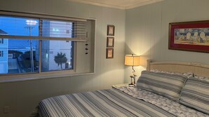 Une chambre à coucher avec un lit, une table de chevet, une lampe, une fenêtre avec des stores et un tableau encadré au mur.