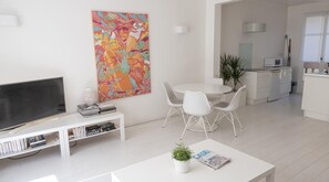 Un salon moderne avec un téléviseur à écran plat, une table basse blanche ornée d’un magazine et d’une plante, un coin repas avec des chaises blanches, et une cuisine en arrière-plan.