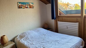 A bedroom with a bed, a nightstand, a decorative lamp, a painting, and a window with a view of houses and trees.