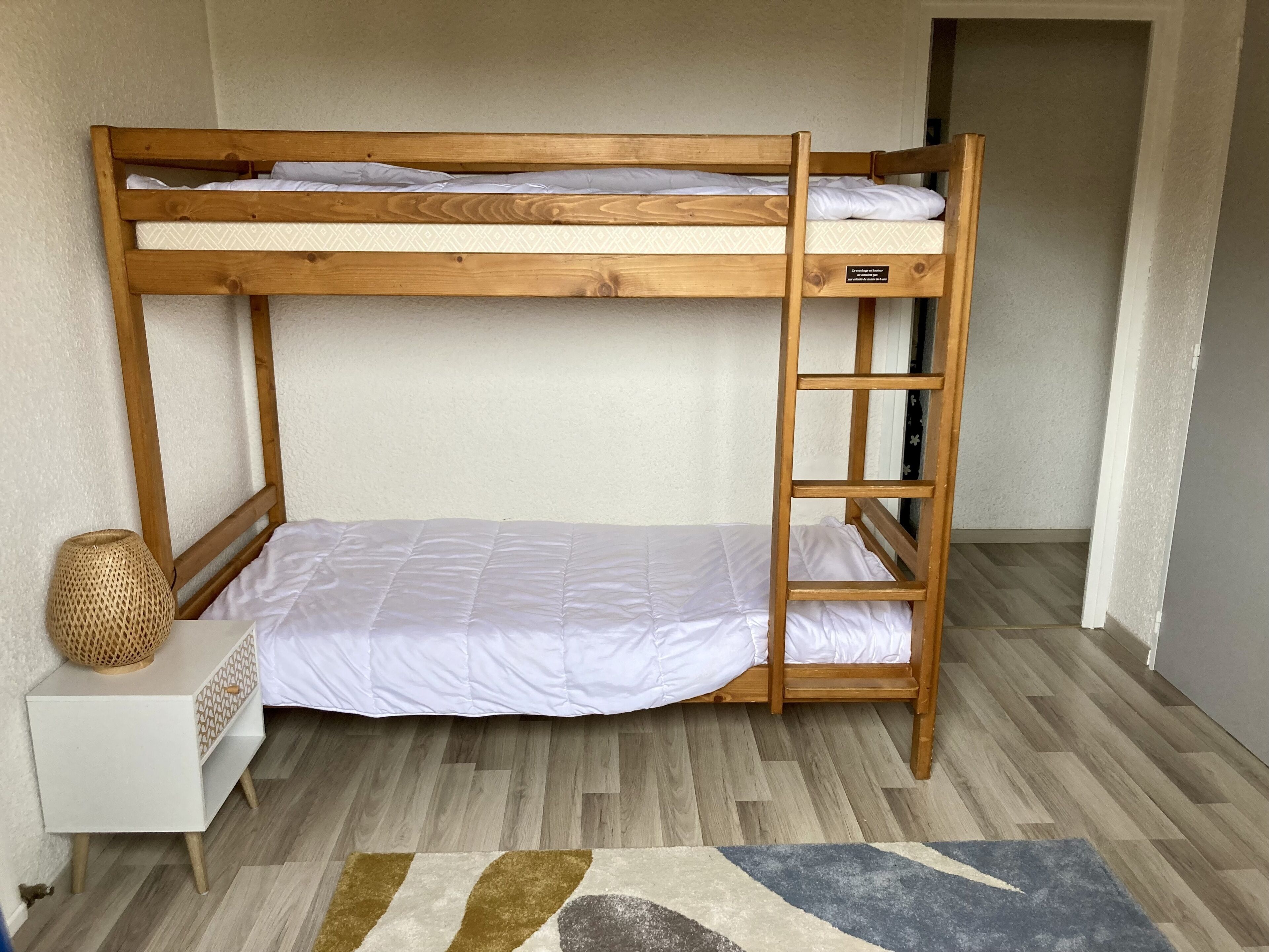 Un lit superposé avec une échelle, une petite table de chevet et un panier en osier.