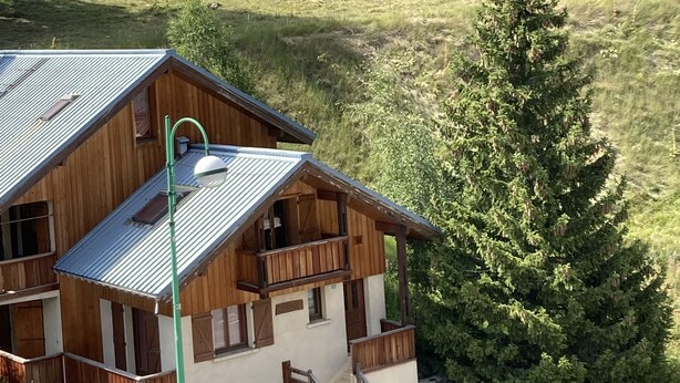 A wooden cabin with a metal roof, a staircase, and a view of a grassy hill with cows.