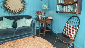 Una habitación con un sofá azul oscuro, un tapete tejido, una mesita de madera con una lámpara, una silla con un cojín estampado y una estantería llena de libros.
