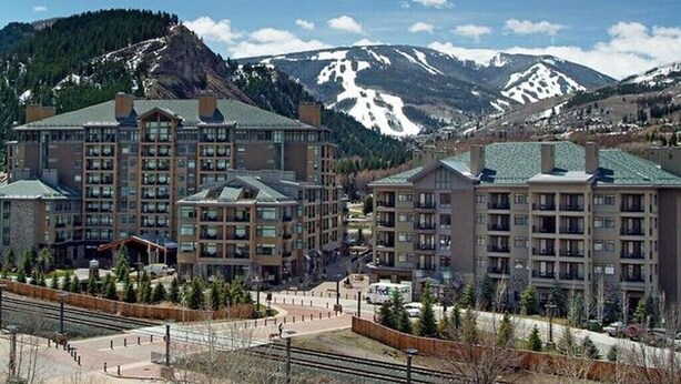 A hotel with multiple buildings, a parking area, and a mountainous backdrop. - Steps from Riverfront Gondola, Modern Cozy Condo, Free Underground Parking (Avon)