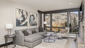 Een moderne woonkamer met een grijze modulaire bank, een ronde salontafel, een fauteuil en een open haard. Grote ramen bieden uitzicht op bomen en heuvels.