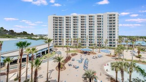 Een hoog hotel met meerdere balkons, een zwembadgedeelte met ligbedden en palmbomen.