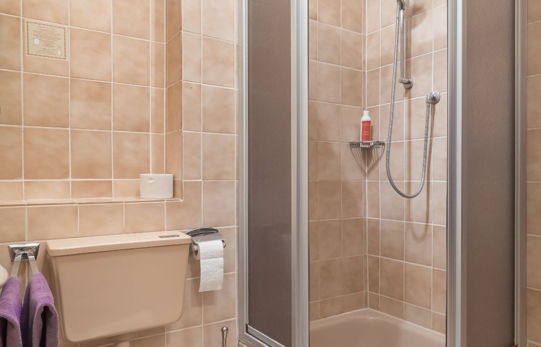 A bathroom with beige tiles, a toilet, a shower with a handheld showerhead, and a towel rack with purple towels.