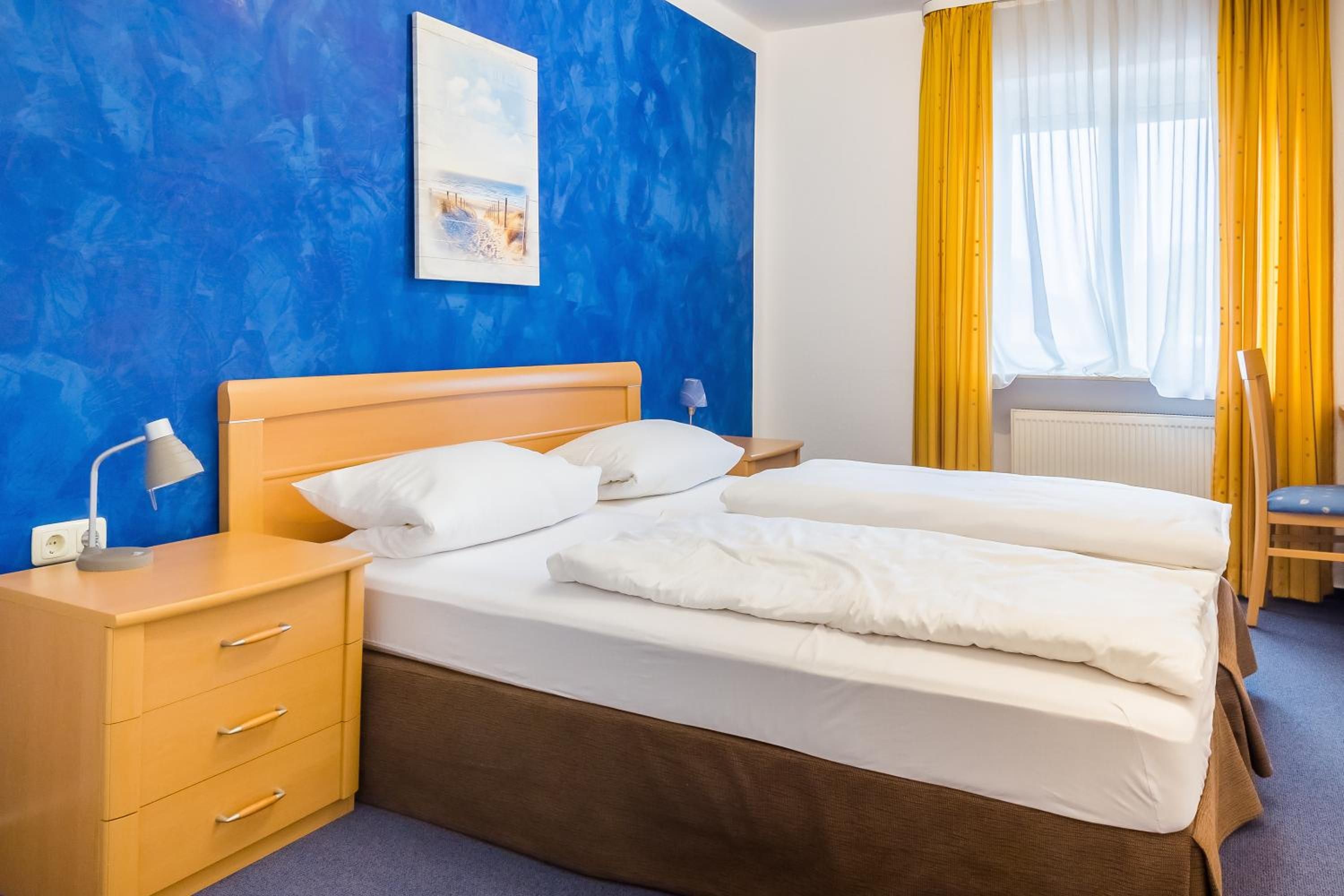 A hotel room with a double bed, a bedside table with a lamp, a chair, and a window with yellow curtains.