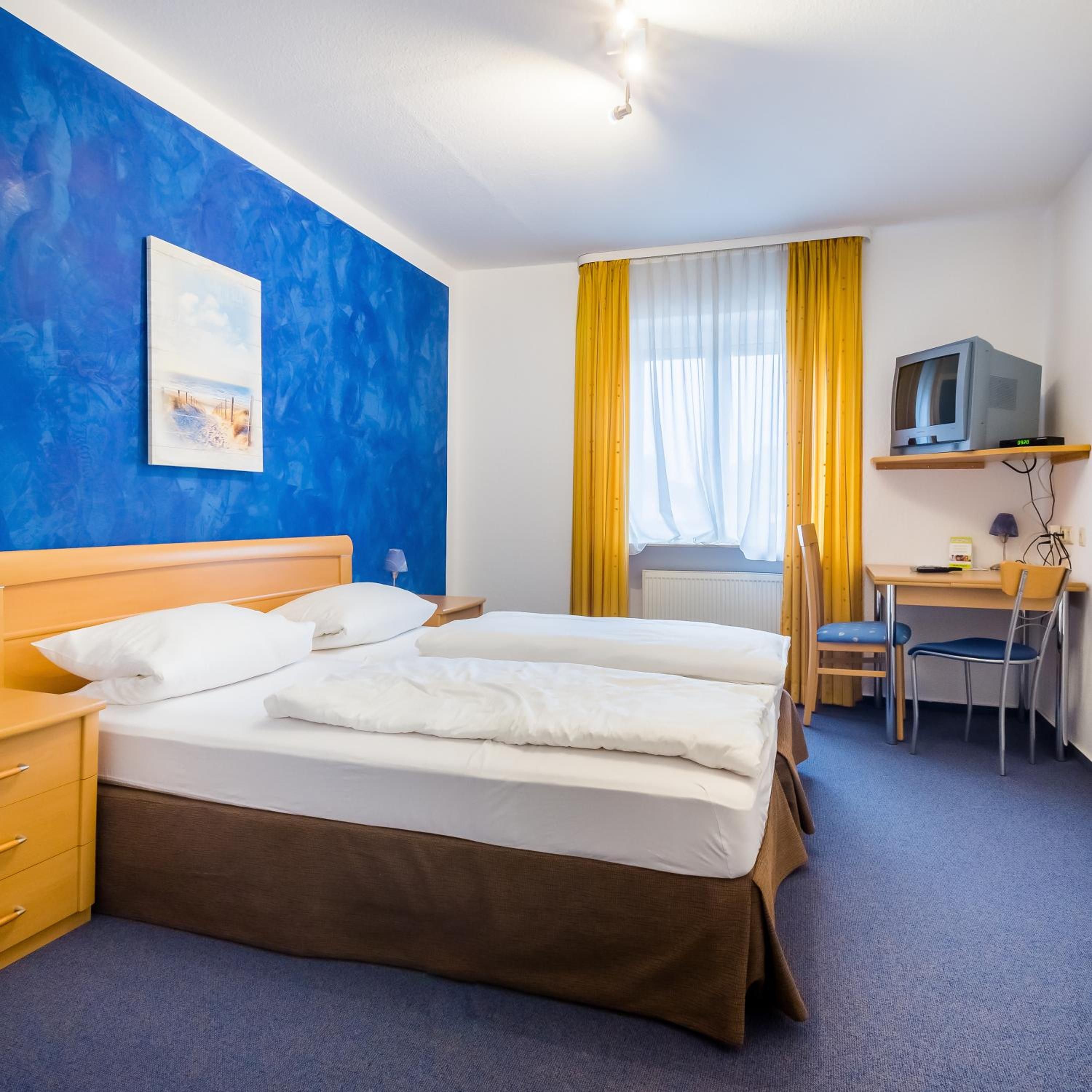 Habitación de hotel con cama doble, escritorio con silla, televisor en un estante y un cuadro en la pared.