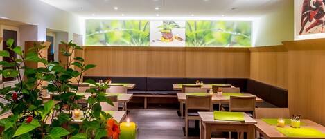 Un restaurante con mesas y bancos de madera, plantas verdes y un mural de temática natural.