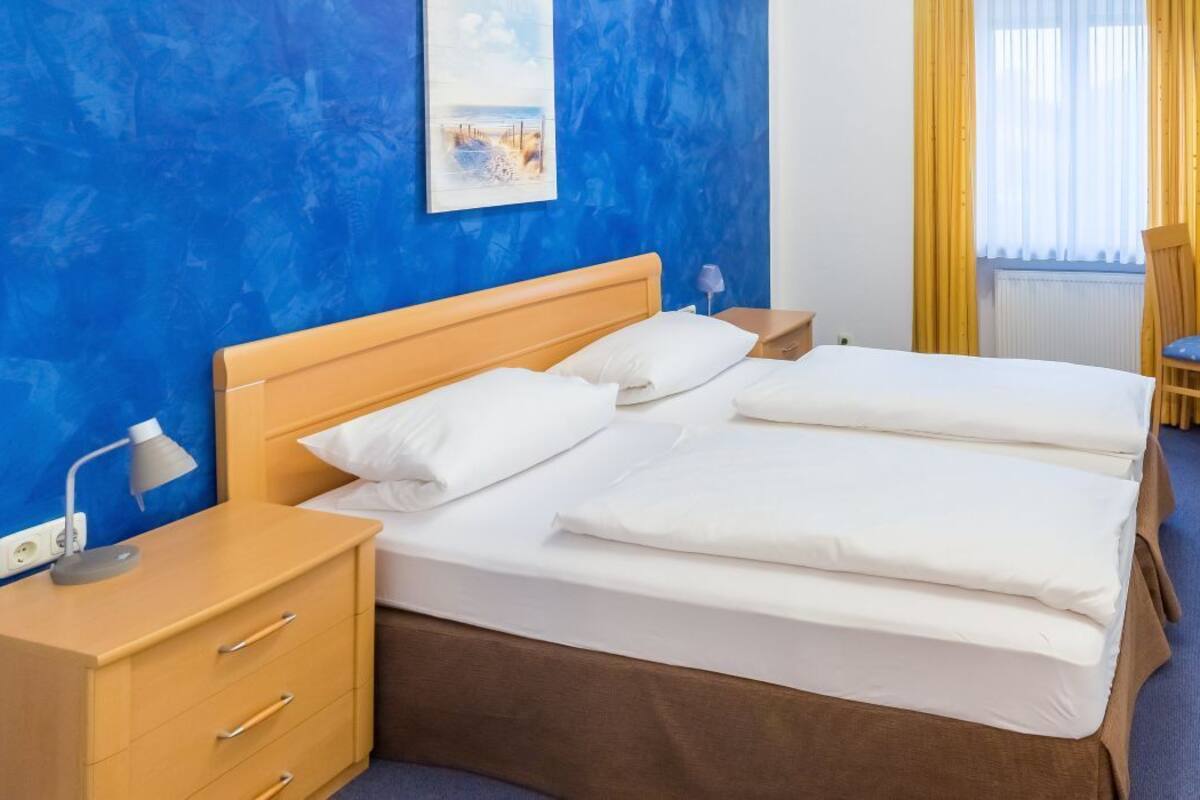 Une chambre d’hôtel avec un lit en bois, une table de chevet avec une lampe, une chaise et une fenêtre avec des rideaux jaunes.