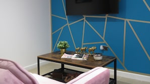 Een kamer met een blauwe geometrische wand, een flatscreen televisie boven een houten salontafel en een roze stoel.