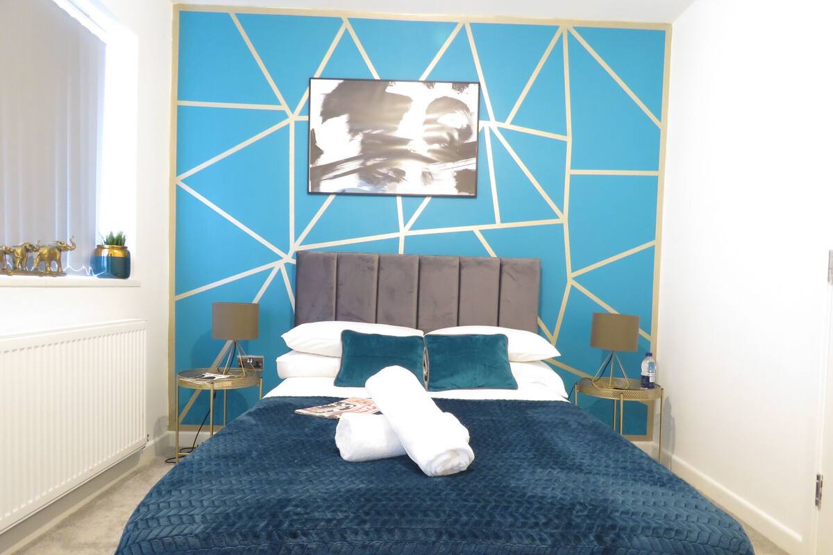 Une chambre à coucher avec un motif géométrique sur un mur, un lit recouvert d’une courtepointe bleue, deux tables de chevet avec des lampes et une fenêtre avec des rideaux.