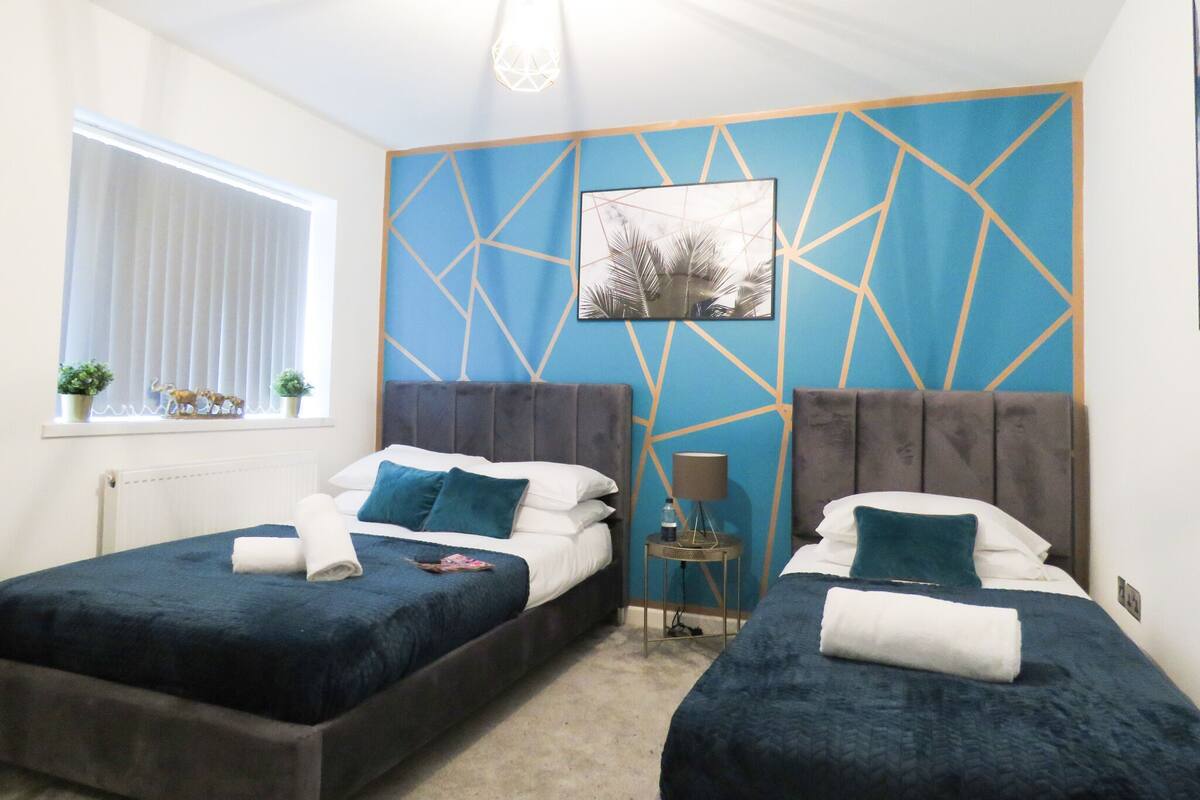 Une chambre avec deux lits, une fenêtre avec des stores, un mur orné d’un motif géométrique et un luminaire au plafond.