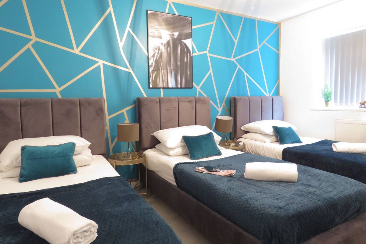 Une chambre d’hôtel avec trois lits, un motif géométrique sur le mur et une grande fenêtre avec des stores.