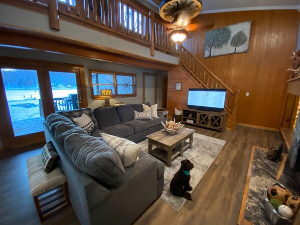 A spacious living room with a large sectional sofa, a television, and a dog sitting on the floor. - Lakefront 4 Bedroom 2.5 Bath Hot Tub, Firepit, Dock, Deck and great backyard! (DuBois)
