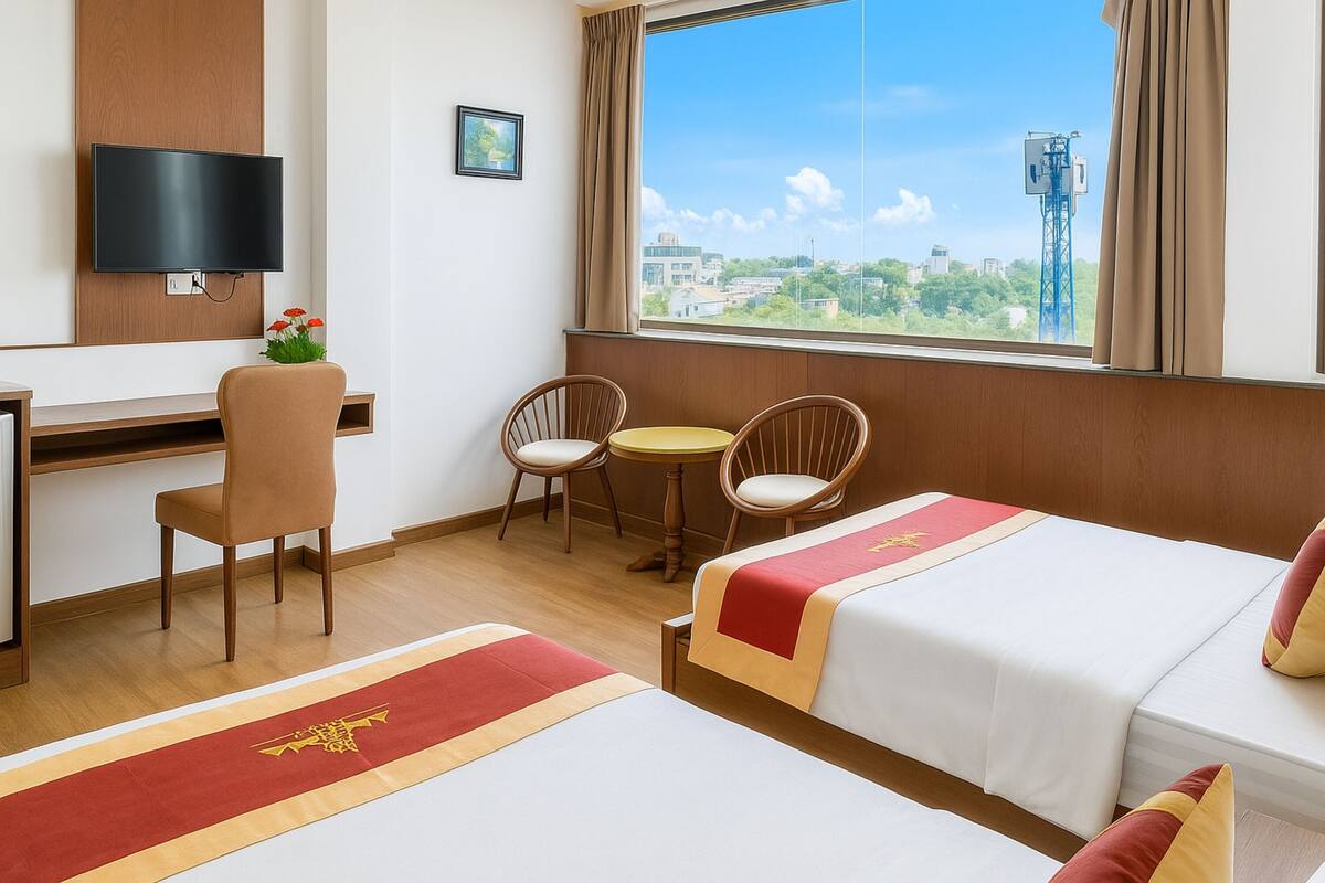 Ein Hotelzimmer mit zwei Betten, einem Schreibtisch, einem Fernseher, einem Fenster mit Stadtblick und einem kleinen Tisch mit Stühlen.