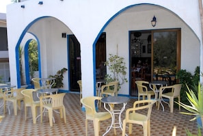 An outdoor patio with yellow plastic chairs and tables, white walls with blue trim, and a tiled floor. - Anthoula Sun (Rhodes)