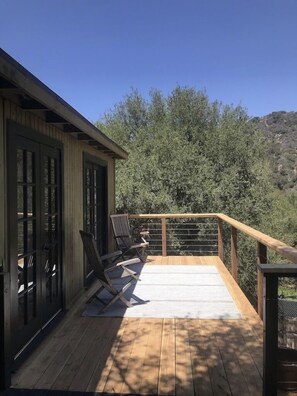 Terraza de madera con barandilla, dos sillas y vista a árboles y colinas.
