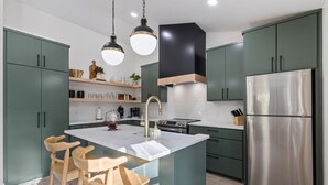 Una cocina moderna con gabinetes verdes, un refrigerador de acero inoxidable, una isla central con taburetes de bar, iluminación colgante y una estufa integrada.