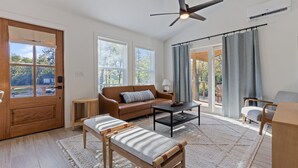 Un salón amplio con un banco de madera, un sofá y una mesa de centro. Hay un ventilador de techo y ventanas grandes que aportan luz natural.