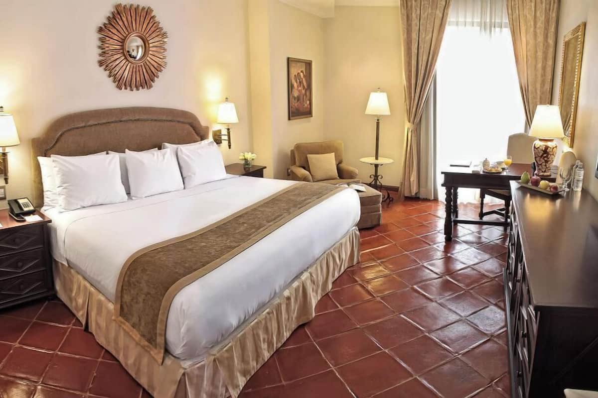 Habitación de hotel con una cama grande, lámparas de noche, un escritorio con lámpara, una silla y un espejo decorativo en la pared.