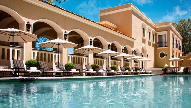 A poolside area with lounge chairs and umbrellas, a clear blue sky, and a building with arches and a tiled roof. - Villa Antigua (Tequisquiapan)