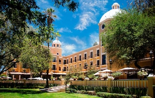 A courtyard with a building featuring a dome and a bell tower, surrounded by lush greenery and outdoor seating areas.